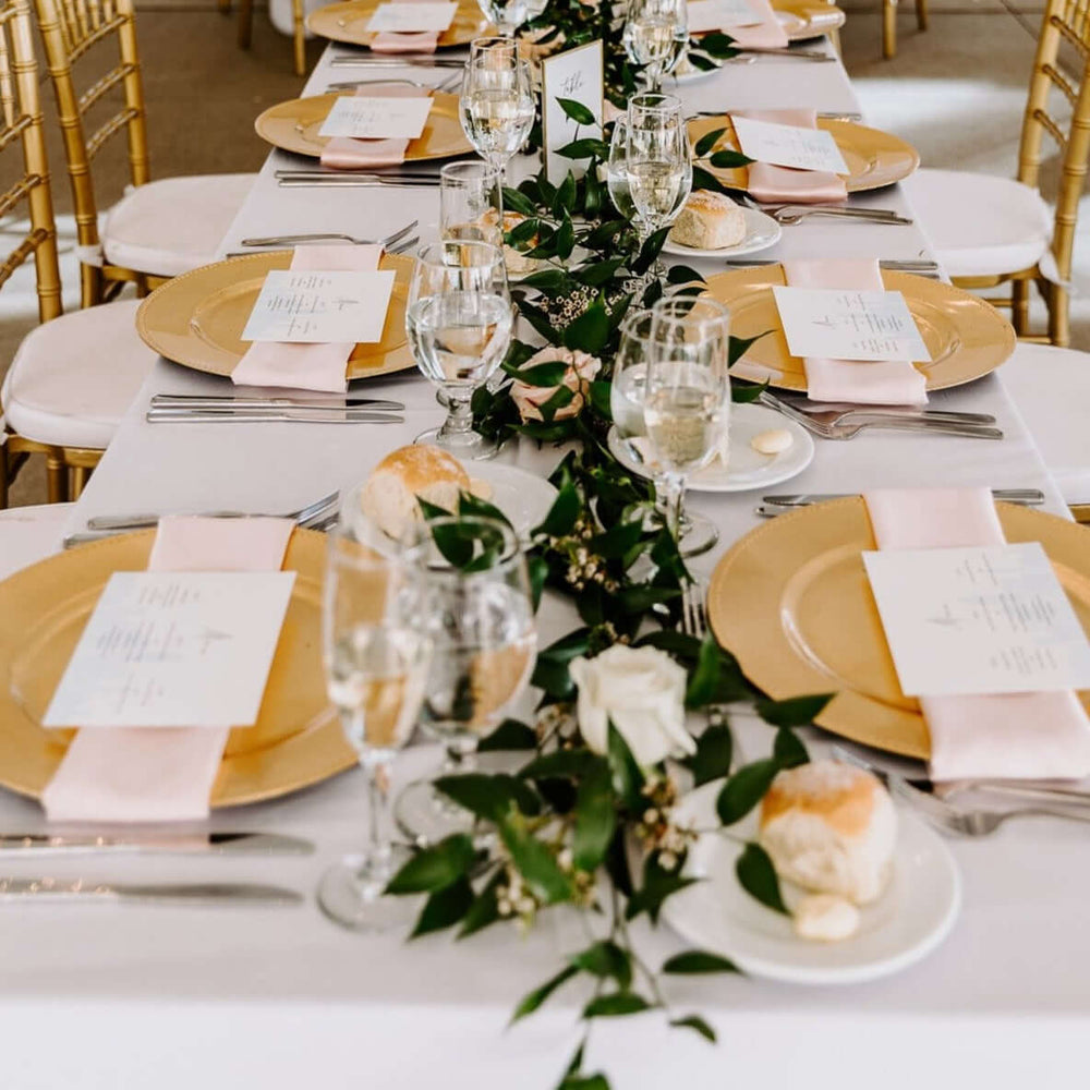 
            
                Load image into Gallery viewer, Fancy table setting with gold chargers and wineglasses with greens along center.
            
        